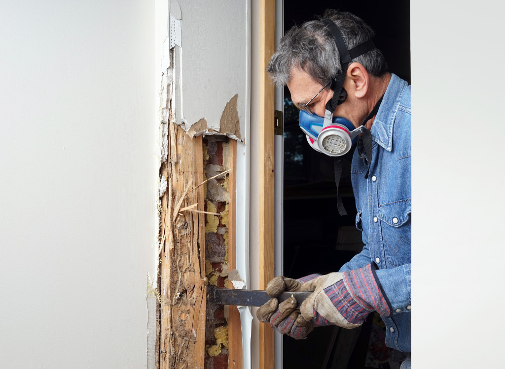 A man wearing a mask removing damaged wood from a wall